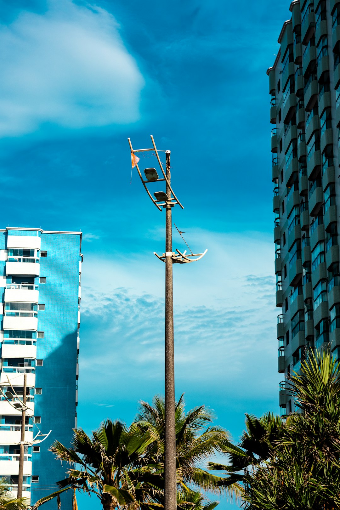 The Art of Drawing Readers In: Your attractive post title goes here A tall, weathered lamppost stands between two modern apartment buildings under a bright blue sky with scattered clouds. Palm trees frame the bottom of the image, adding a tropical feel to the urban landscape.